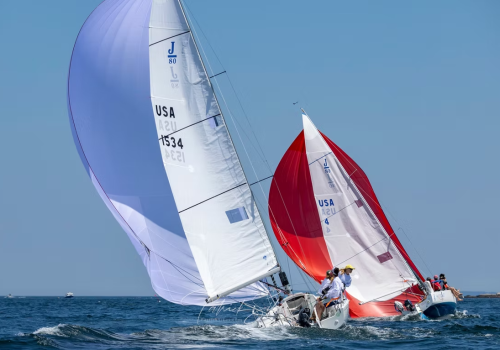 Two sailboats race on the open water with colorful sails, one white and one red, under a clear blue sky.