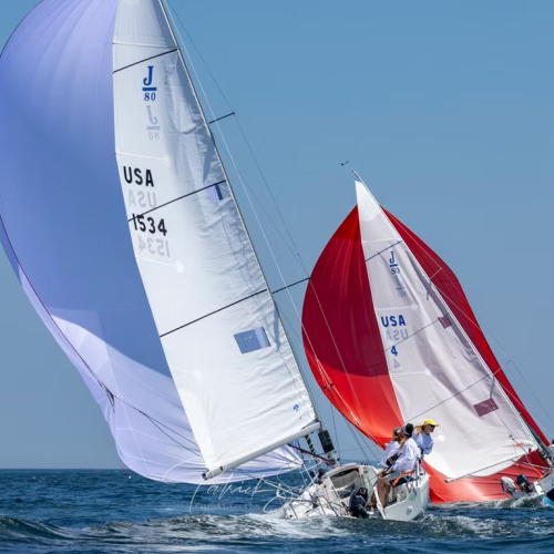 Two sailboats race on the open water with colorful sails, one white and one red, under a clear blue sky.