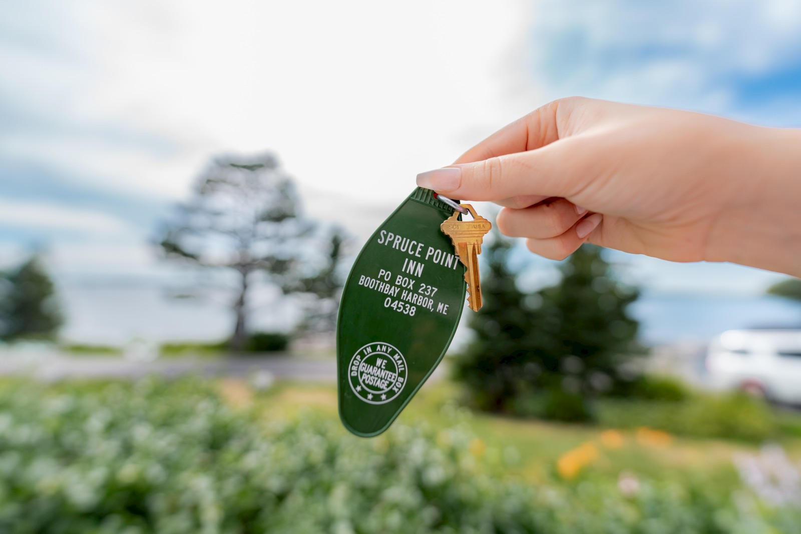 A hand holds a green hotel keychain for Spruce Point Inn in Boothbay Harbor, ME, with keys attached against a blurred outdoor background.