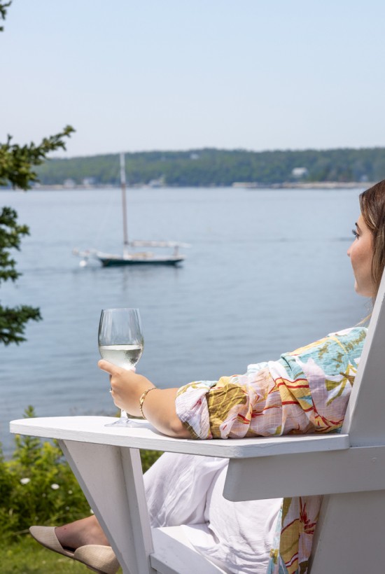 A person sits in a white chair holding a glass, relaxing by a calm body of water with a sailboat in the distance and a tree nearby.