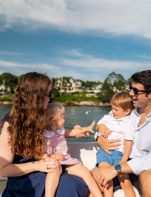 A family enjoys a sunny day on a boat, seated together, with water and houses visible in the background under a partly cloudy sky.