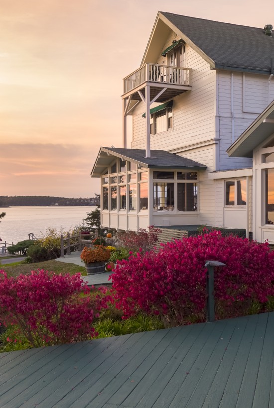 A house with large windows overlooks a scenic waterfront at sunset, surrounded by vibrant plants and trees, with a wooden deck in the foreground.