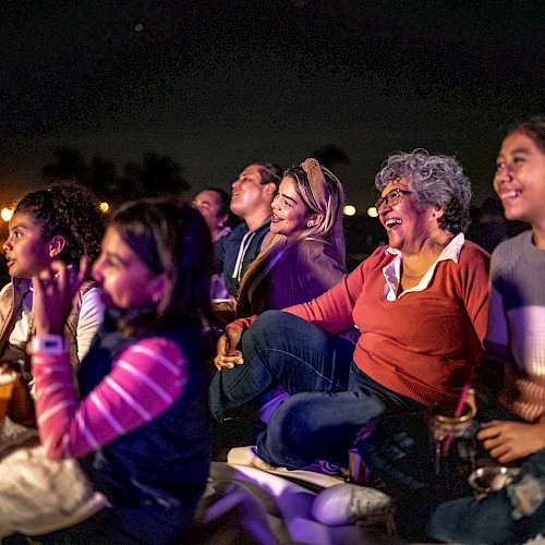 People sitting outdoors, smiling and watching something at night, with buckets of popcorn.