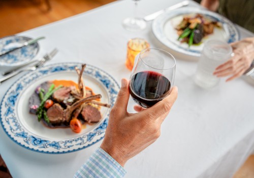 A table setting featuring two plates of lamb chops with vegetables, a hand holding a glass of red wine, and another holding a glass of water.