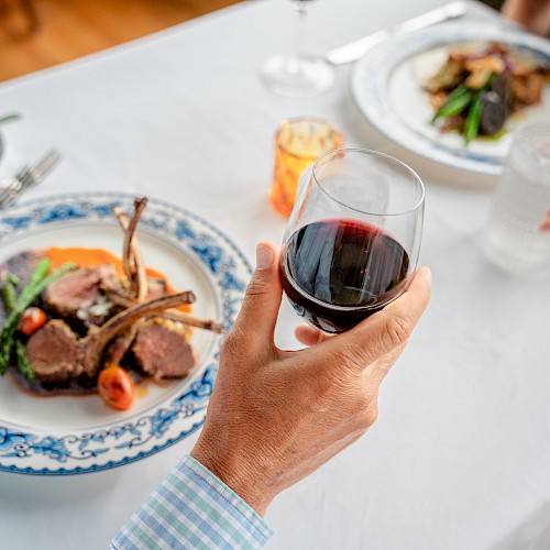 A table setting featuring two plates of lamb chops with vegetables, a hand holding a glass of red wine, and another holding a glass of water.