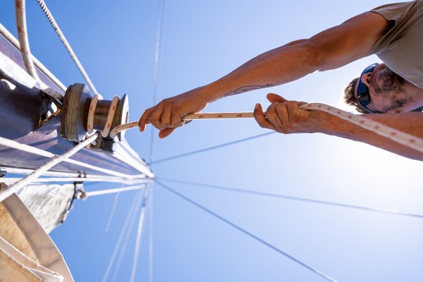 A person on a sailboat adjusts a rope against a clear blue sky, with sunlight highlighting the scene and rigging visible in the background.