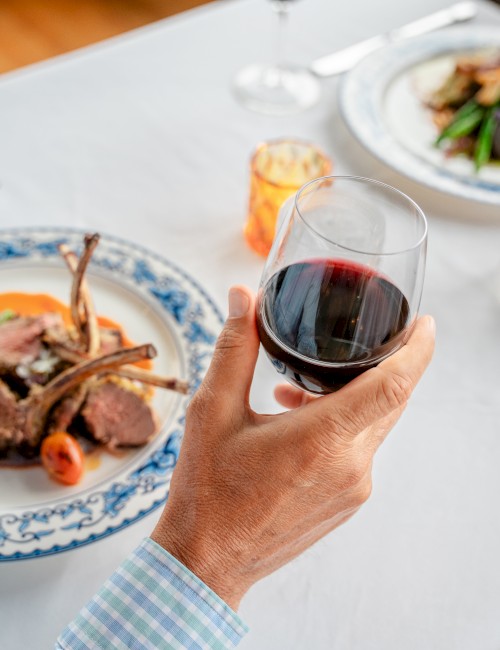 Two plates with lamb chops and vegetables on a table; a hand holds a glass of red wine. Another hand with a water glass is visible.