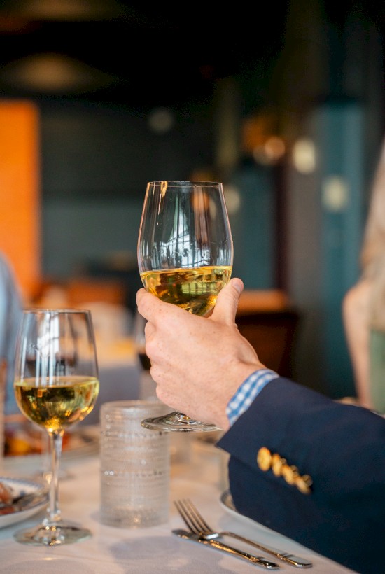 A person holding a glass of white wine in a restaurant setting, with another glass and dining utensils visible on the table.