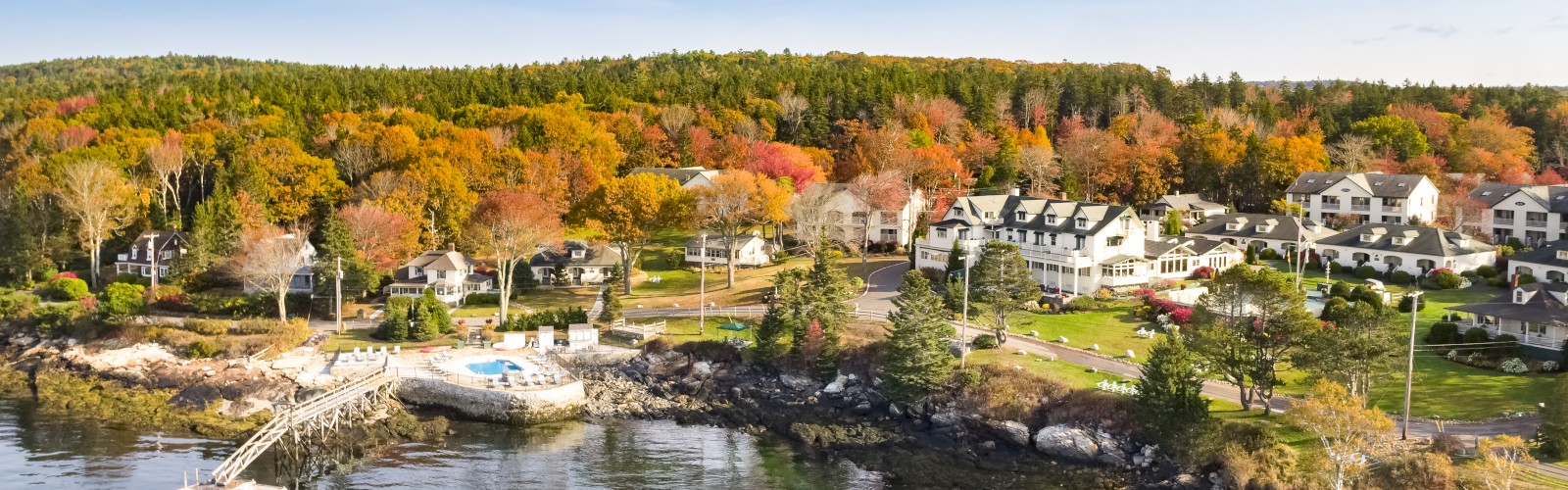 A coastal village with houses, autumn-colored trees, and a dock on calm water under a clear sky.