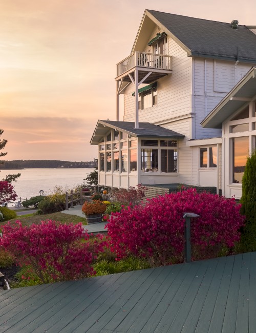A house with large windows overlooks a scenic waterfront at sunset, surrounded by vibrant plants and trees, with a wooden deck in the foreground.