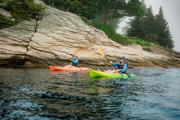Two people are kayaking near a rocky shoreline with trees in the background, one in an orange kayak and the other in a green kayak.
