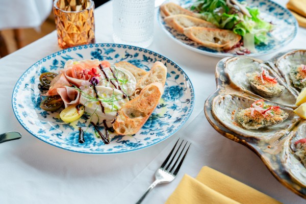 A plate with charcuterie and bread, another with oysters, plus a drink and salad on the table, set on a white tablecloth.