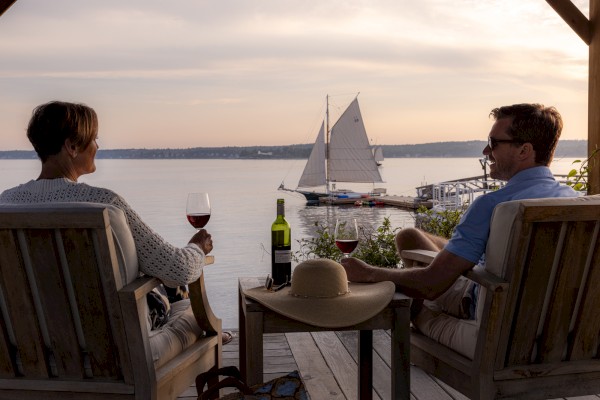 Two people relax with wine on a deck, overlooking a calm body of water as a sailboat passes by under a golden sky.