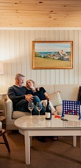 A couple sits on a sofa in a cozy living room with a board game, wine bottle, and glasses on the table, under a framed painting.