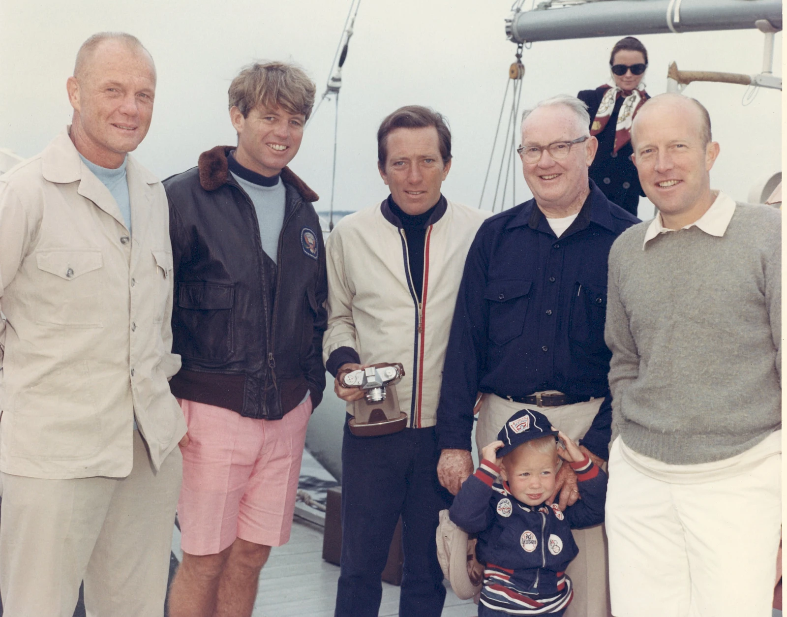 A group of six adults and a child pose on a boat. They're dressed casually, and one person is holding a camera.