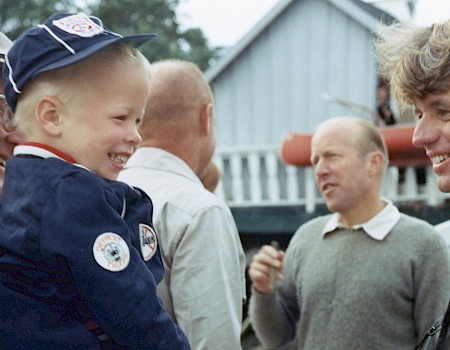 A man in a black jacket smiles, interacting with an older man holding a young boy, with others in the background.