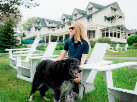 A person in sunglasses sits on a white chair outside, with a large dog standing nearby, and a large house in the background.