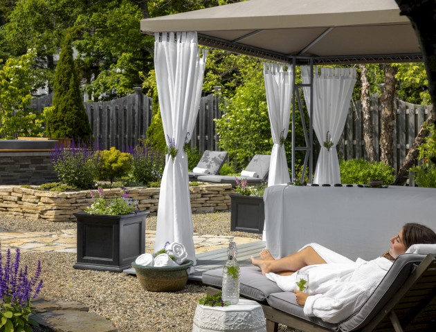 A person relaxes on a lounge chair under a canopy in a garden with potted plants, flowers, and a stone walkway.