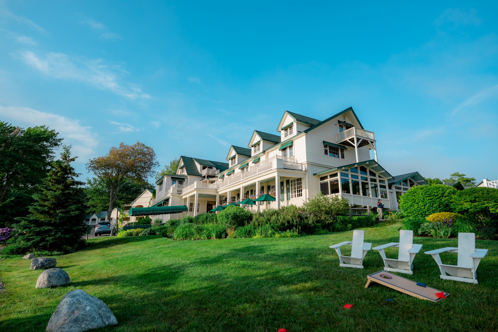 A large house with green roofs sits on a grassy hill, surrounded by trees and Adirondack chairs. A blue sky stretches overhead.