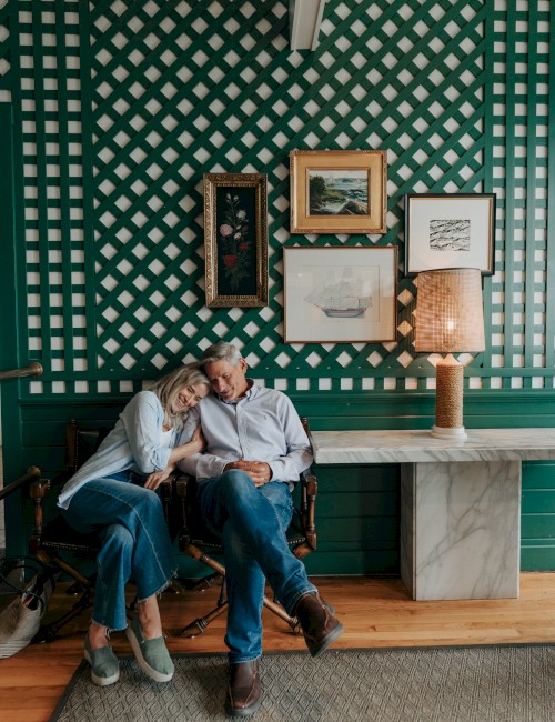 A couple sits closely in a cozy room with a green checkered wall, framed art, and a lamp on a stone table.