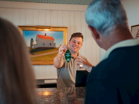 A bartender pours green liqueur for two customers at a bar, inside a cozy room with framed artwork on the wall, smiling gesture.