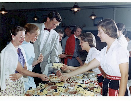 A crowded bake sale or reception with people serving cake to a line of customers, vintage vibe and uniforms.