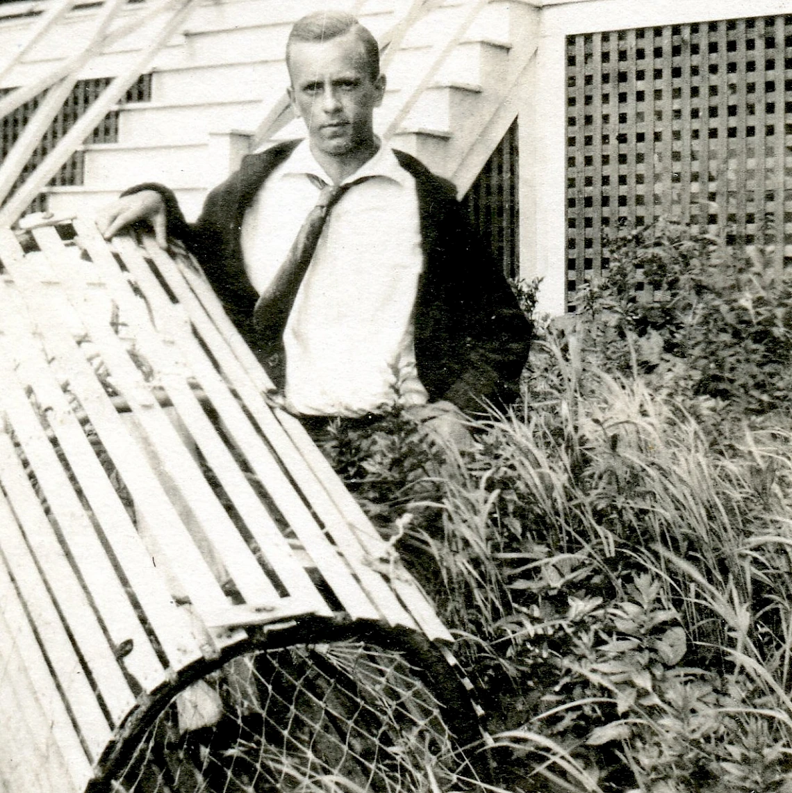 A man in suspenders stands on a porch with a broken fence and overgrown plants, gripping a railing and looking toward the camera.