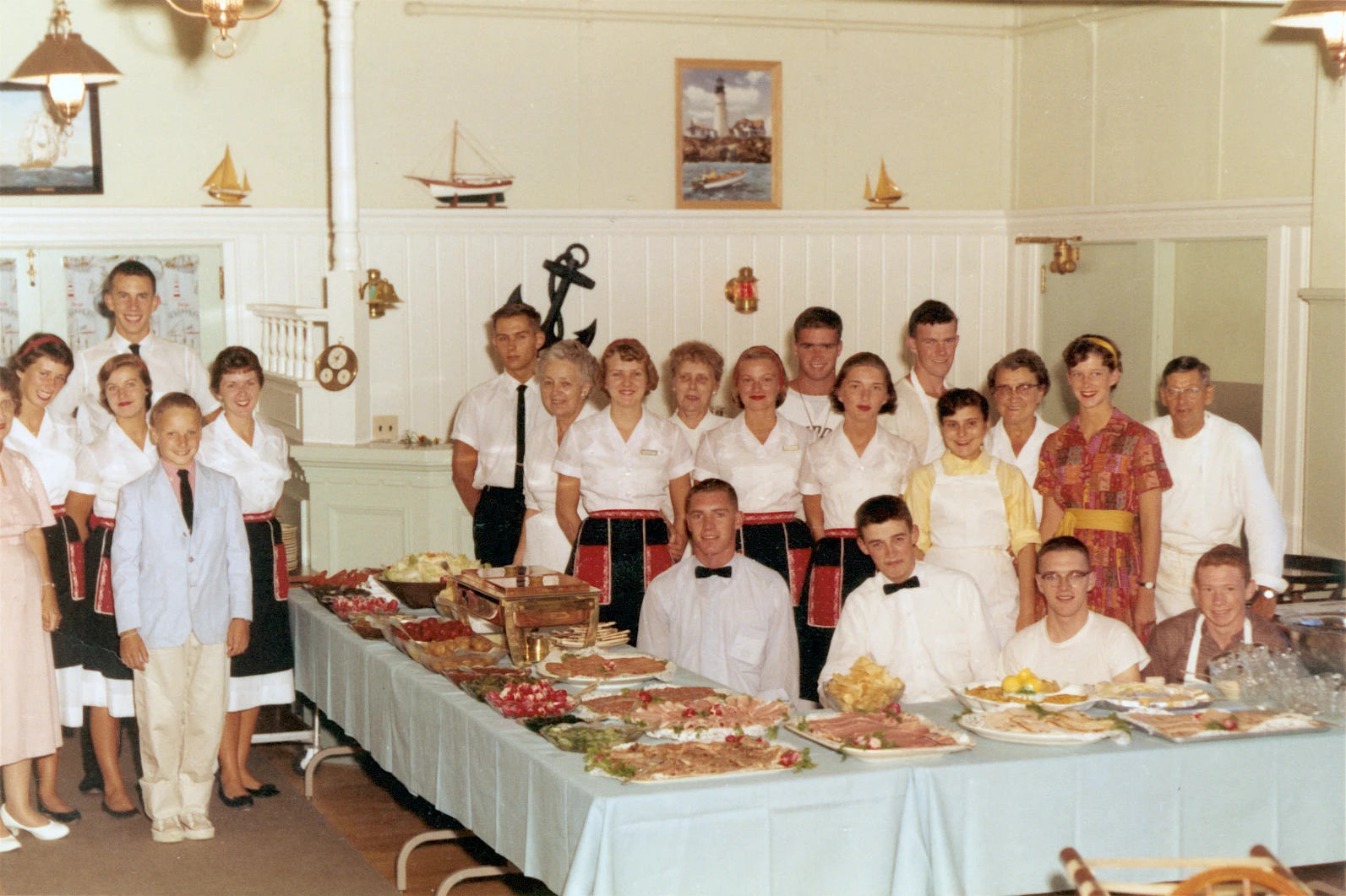 A group of staff posing behind a long banquet table with a variety of dishes, all dressed in white shirts; a vintage restaurant or event setting.