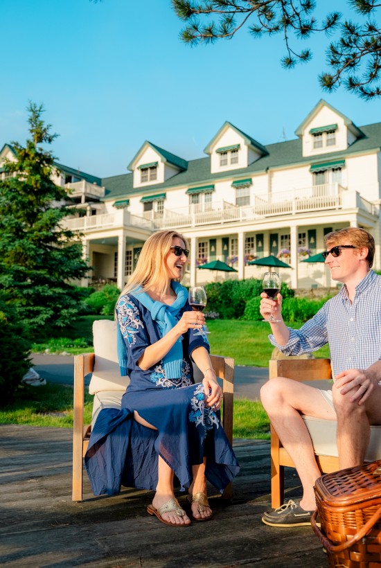 Two people sit on a wooden deck having a sunny chat with a large house and trees in the background, a picnic basket nearby on a bright day.