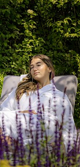 A woman relaxing outdoors in a white robe on a lounge chair among lavender and greenery, with a bottle and glass on a small table.