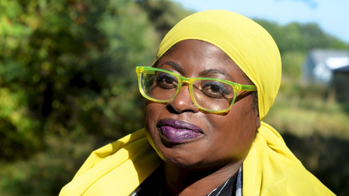 A woman wearing a yellow headwrap and glasses outdoors on a sunny day.