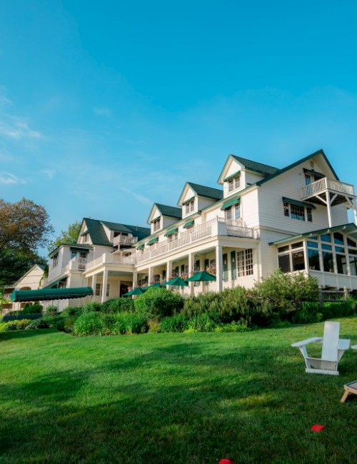A large seaside resort with white buildings, a green lawn, and white Adirondack chairs on the grass under a bright blue sky.