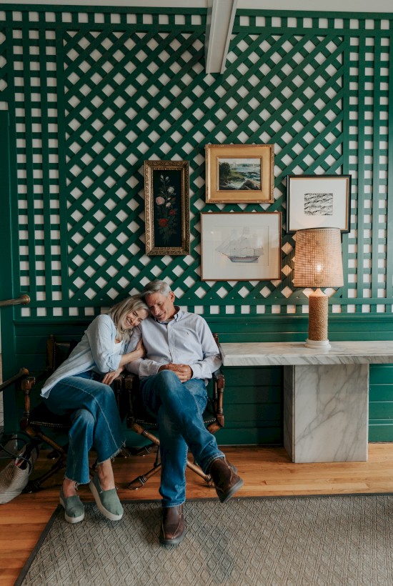 Two people lean close, smiling, seated on a bench in a green patterned room with framed art, a lamp, and a wooden floor.
