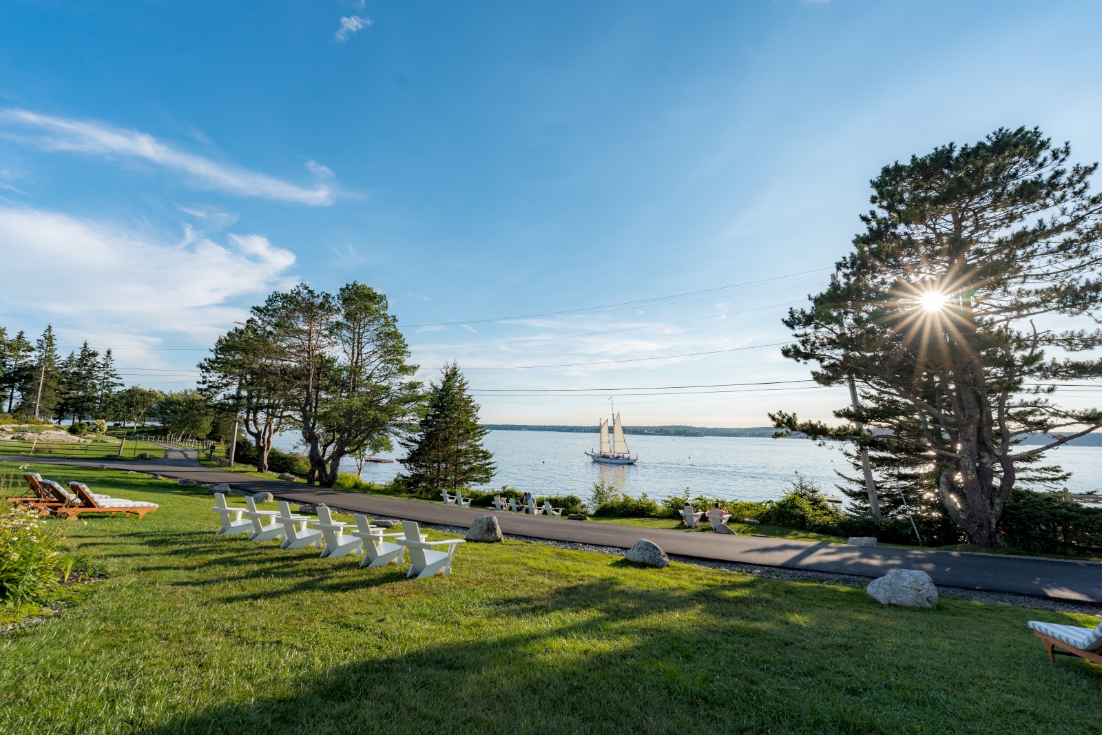 A scenic view of a park with chairs on the grass, a road, trees, and a sailboat on a large body of water beneath a sunny, blue sky.