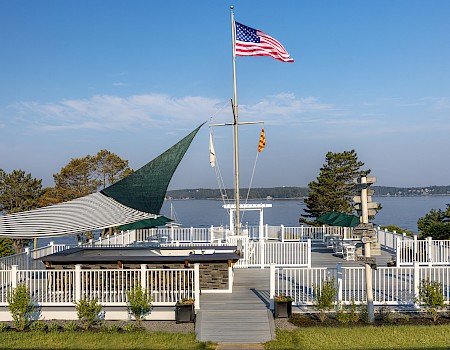 A scenic lakeside patio with white fencing, flags, outdoor seating, and a view of the water under a blue sky.