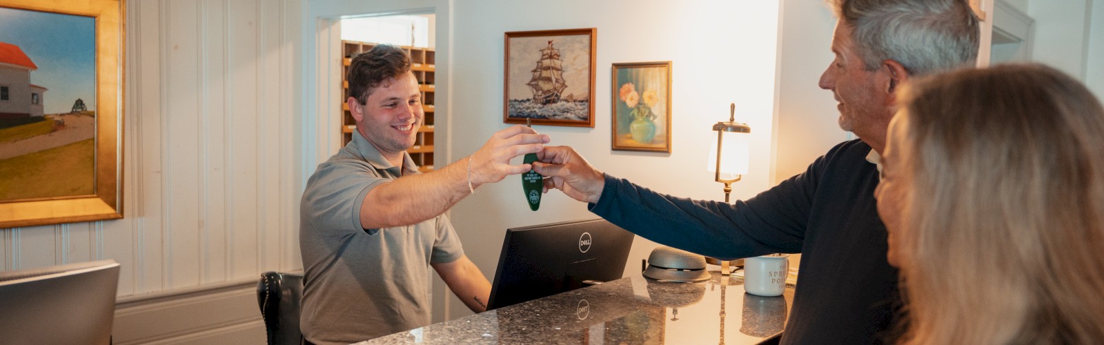 A young man at a reception desk hands key fob to an older man, with a woman beside him, in a cozy, decorated indoor setting.
