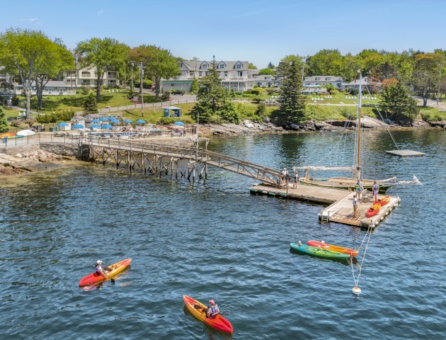 A small marina with people kayaking, a sailboat docked, and a lush green background under a clear blue sky.