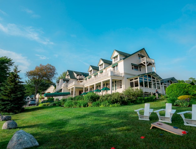 A large, white, multi-story house with green roofs, surrounded by greenery, outdoor chairs, and a wood board game on the lawn.