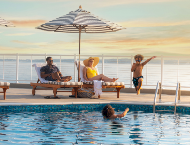 People relaxing and children playing on a poolside deck with umbrellas, lounge chairs, and a pool overlooking a body of water at sunset.