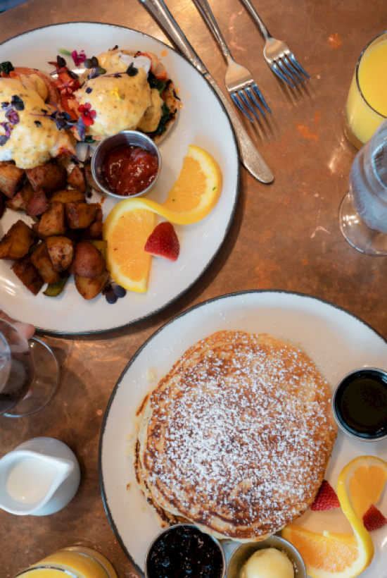 A breakfast includes scrambled eggs with toppings, roasted potatoes, fruits, pancakes with syrup, and various drinks.