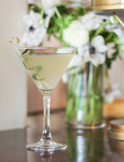 A classic cocktail garnished with an olive on a cocktail pick, placed on a dark surface near a flower arrangement and a gold lamp.